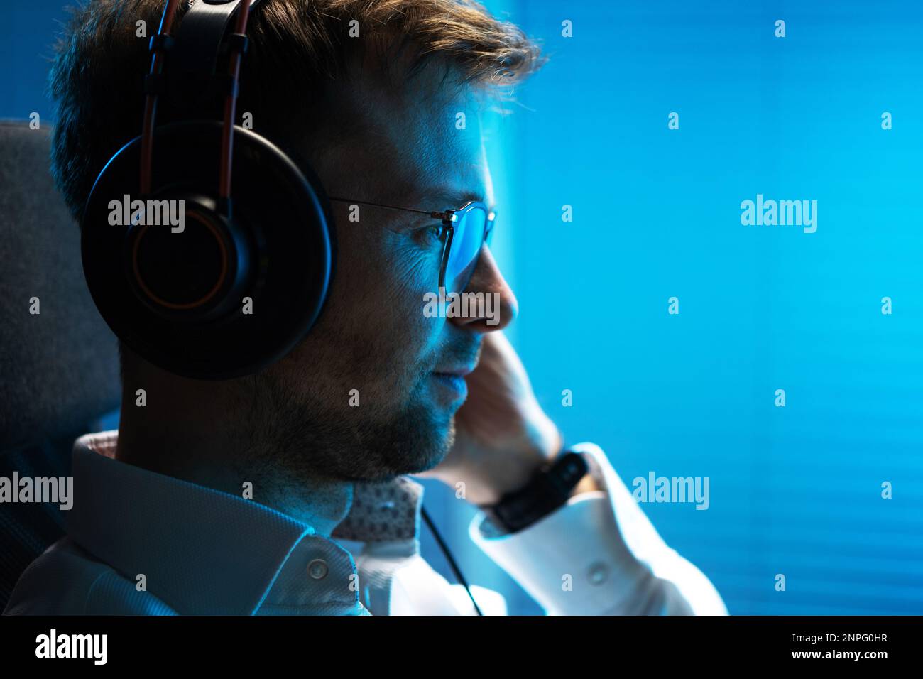 L'uomo caucasico nel suo podcast di ascolto del 30s sul suo computer utilizzando cuffie wireless professionali. Scena di illuminazione blu scuro. Foto Stock