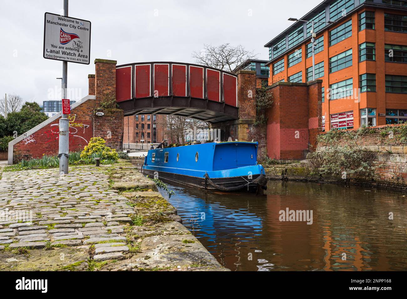 Una barca stretta blu passa sotto il grazioso ponte pedonale rosso Kitty nella zona degli ancoats di Manchester nel 2023. Foto Stock