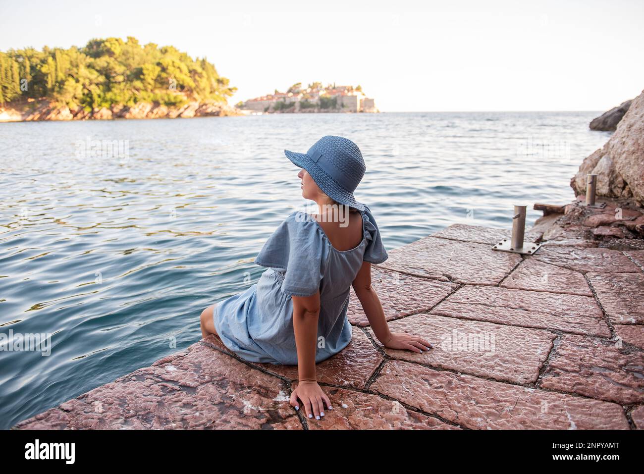 Donna incinta senza volto in abito denim, cappello di paglia blu siede sul mare, guarda l'isola di Sveti Stefan in Montenegro al tramonto. Turismo, viaggi Foto Stock