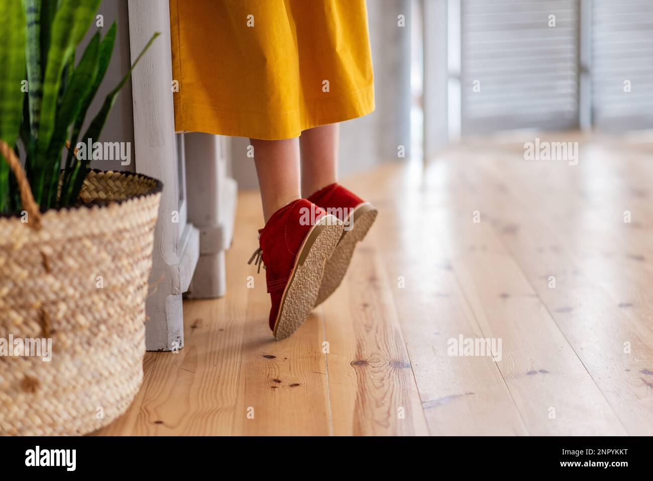 Ragazza in abito giallo profondo è in piedi sul tiptoe da armadio in legno in cucina. Scarpe per bambini in borgogna da primo piano su un pavimento in laminato chiaro. Foto Stock
