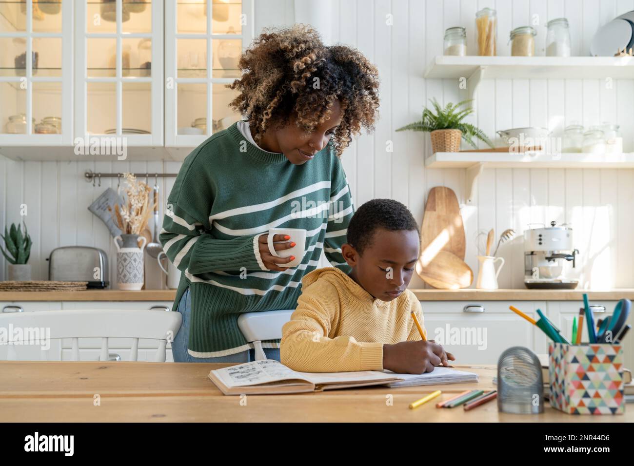 Mamma premurosa donna di razza mista con la tazza guardare il figlio portatile di scrittura a mano. Homeschooling insieme genitore Foto Stock