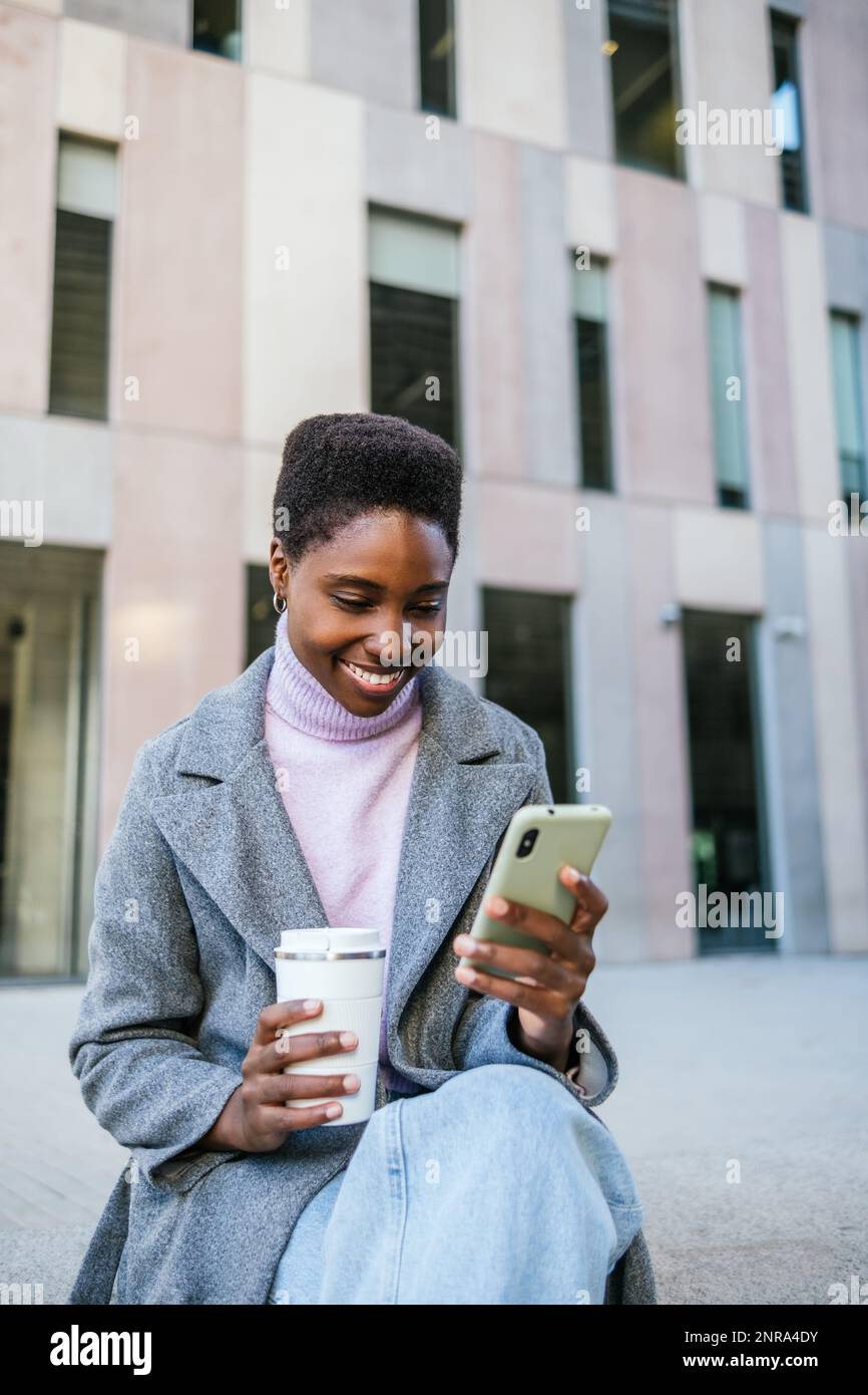 Allegra giovane donna nera in elegante outerwear con tazza di caffè ecocompatibile. Sorride e naviga sui social media sul telefono cellulare mentre siede sulla panchina di fronte a un edificio moderno sulla strada cittadina Foto Stock