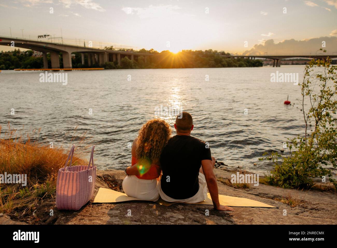 Vista posteriore della coppia guardando il tramonto mentre si siede su coperta da picnic vicino al mare Foto Stock