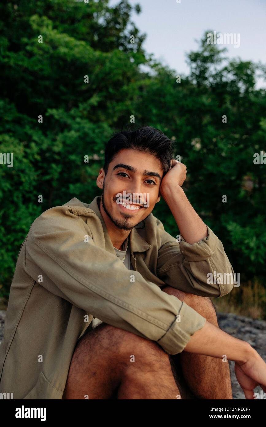 Ritratto di un giovane sorridente che appoggia la testa sul gomito mentre si siede contro le piante Foto Stock