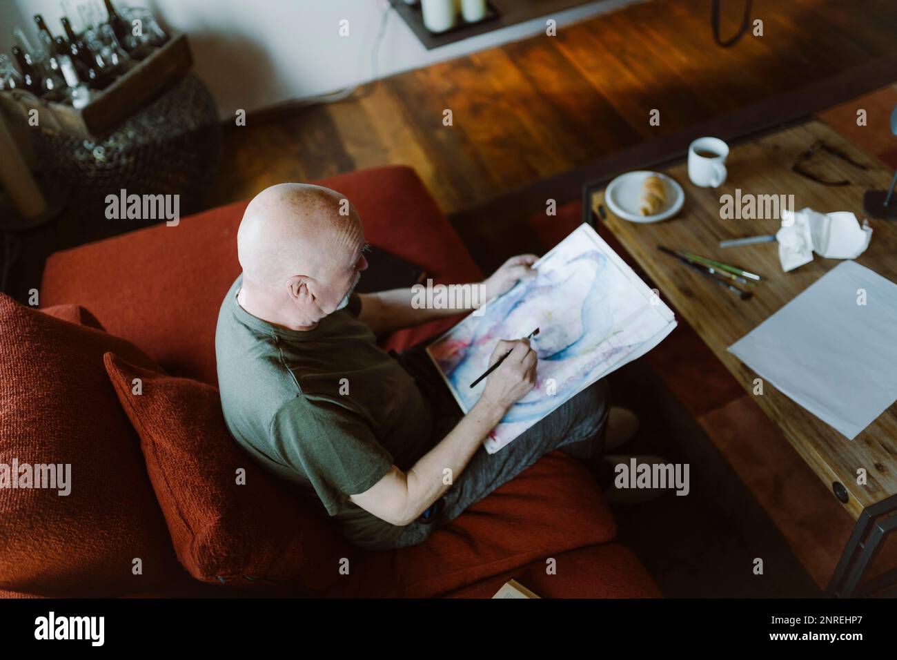 Vista dall'alto dell'uomo anziano che dipinge la faccia sopra la carta mentre si siede sul divano a casa Foto Stock