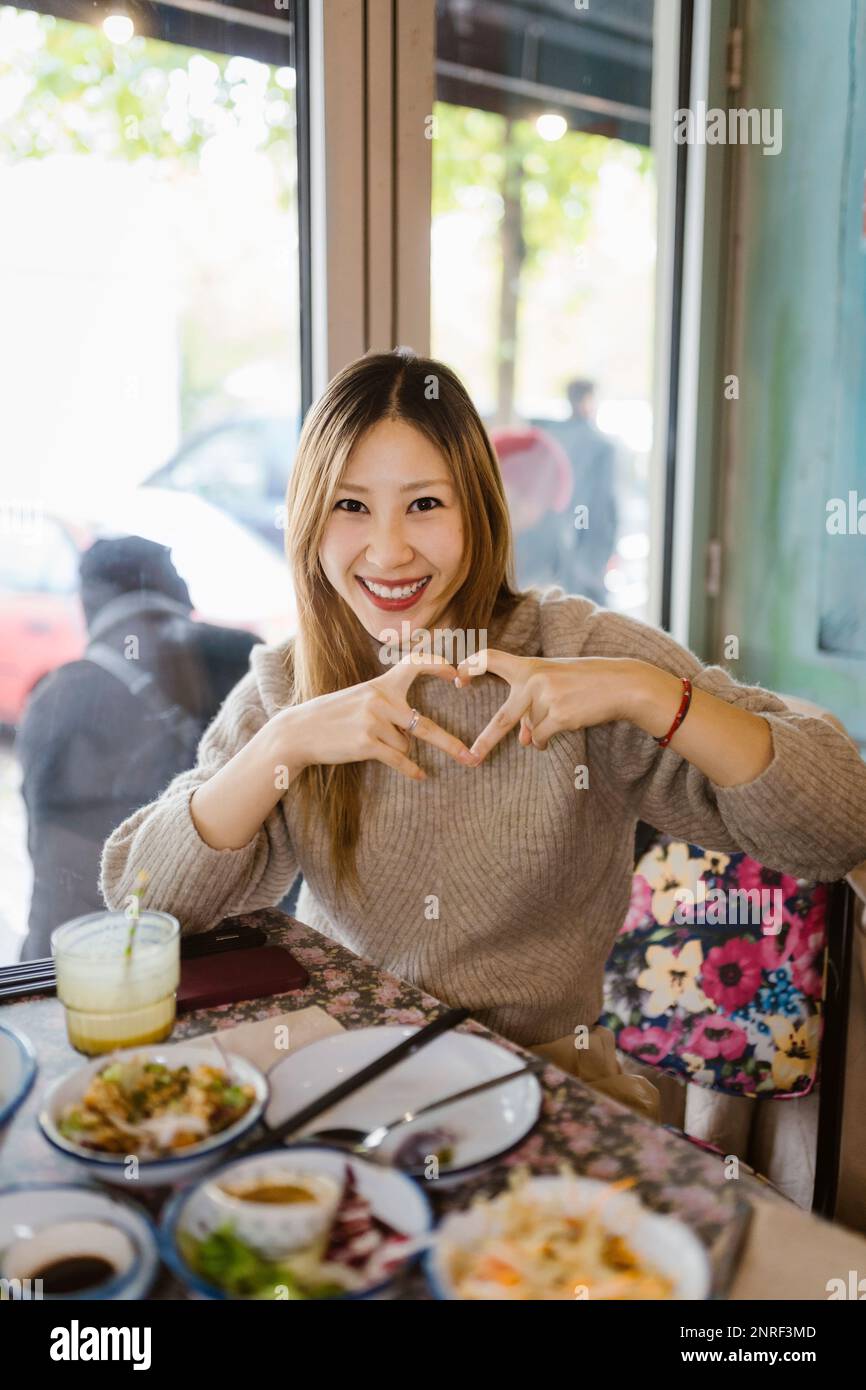 Ritratto di donna felice facendo gesto a forma di cuore mentre si siede al ristorante Foto Stock