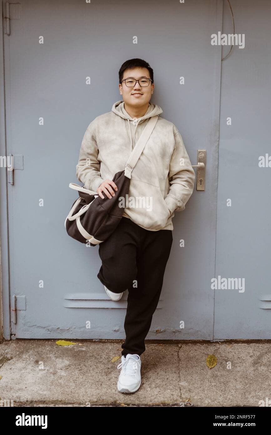 Giovane uomo sorridente con la mano in tasca appoggiata sulla porta grigia Foto Stock