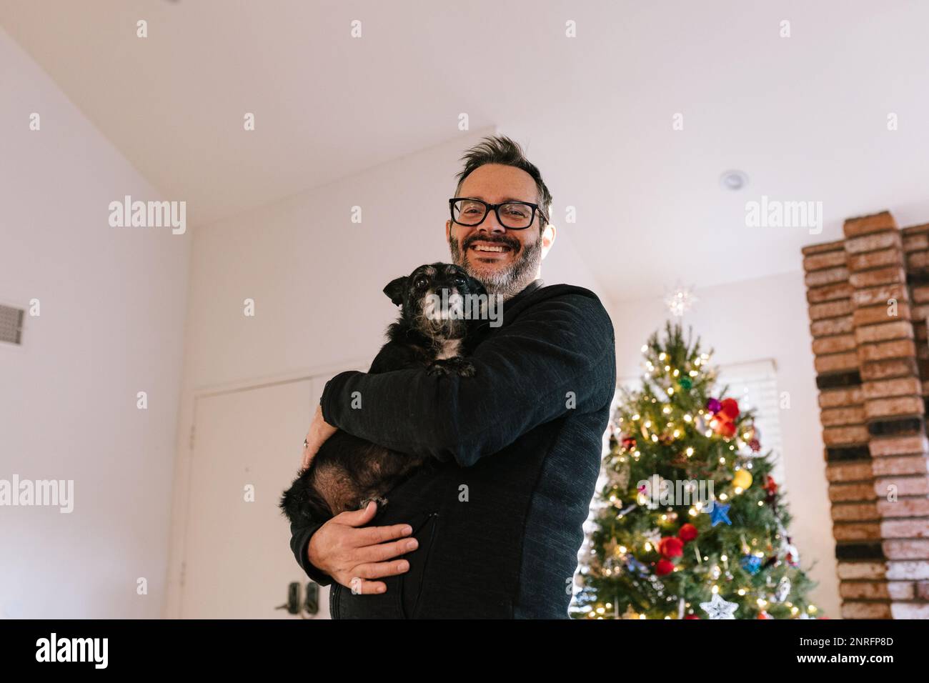 Uomo felice che tiene un piccolo cane nero vicino all'albero di natale Foto Stock