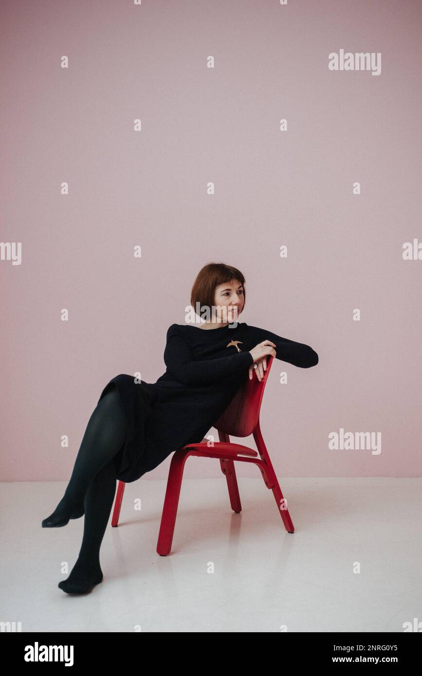 una donna in un vestito nero siede su una sedia, sfondo rosa Foto Stock