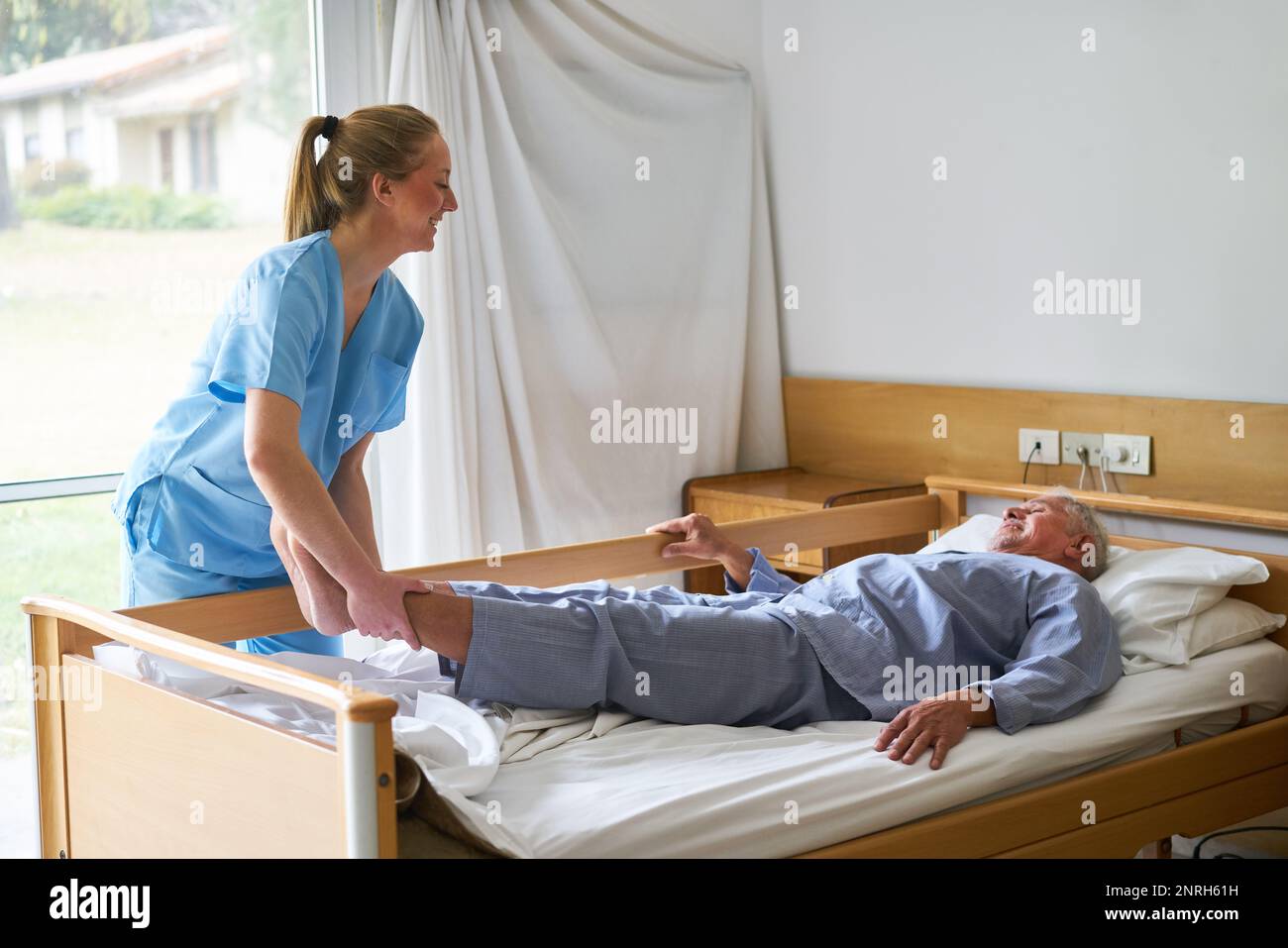 Giovane infermiera sorridente che solleva le gambe dell'uomo anziano sdraiato a letto nella casa di cura Foto Stock