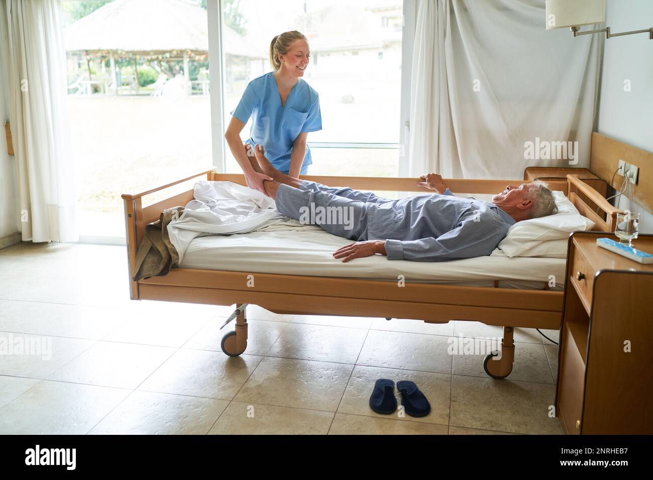 Sorridente infermiera che solleva le gambe di un anziano sdraiato nel letto di casa di cura Foto Stock