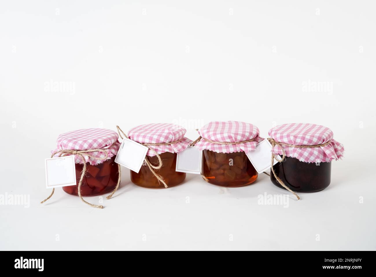 Una serie di vasetti per marmellata con coperchi a scacchi rosa e bianchi ed etichette vuote isolate su sfondo bianco Foto Stock