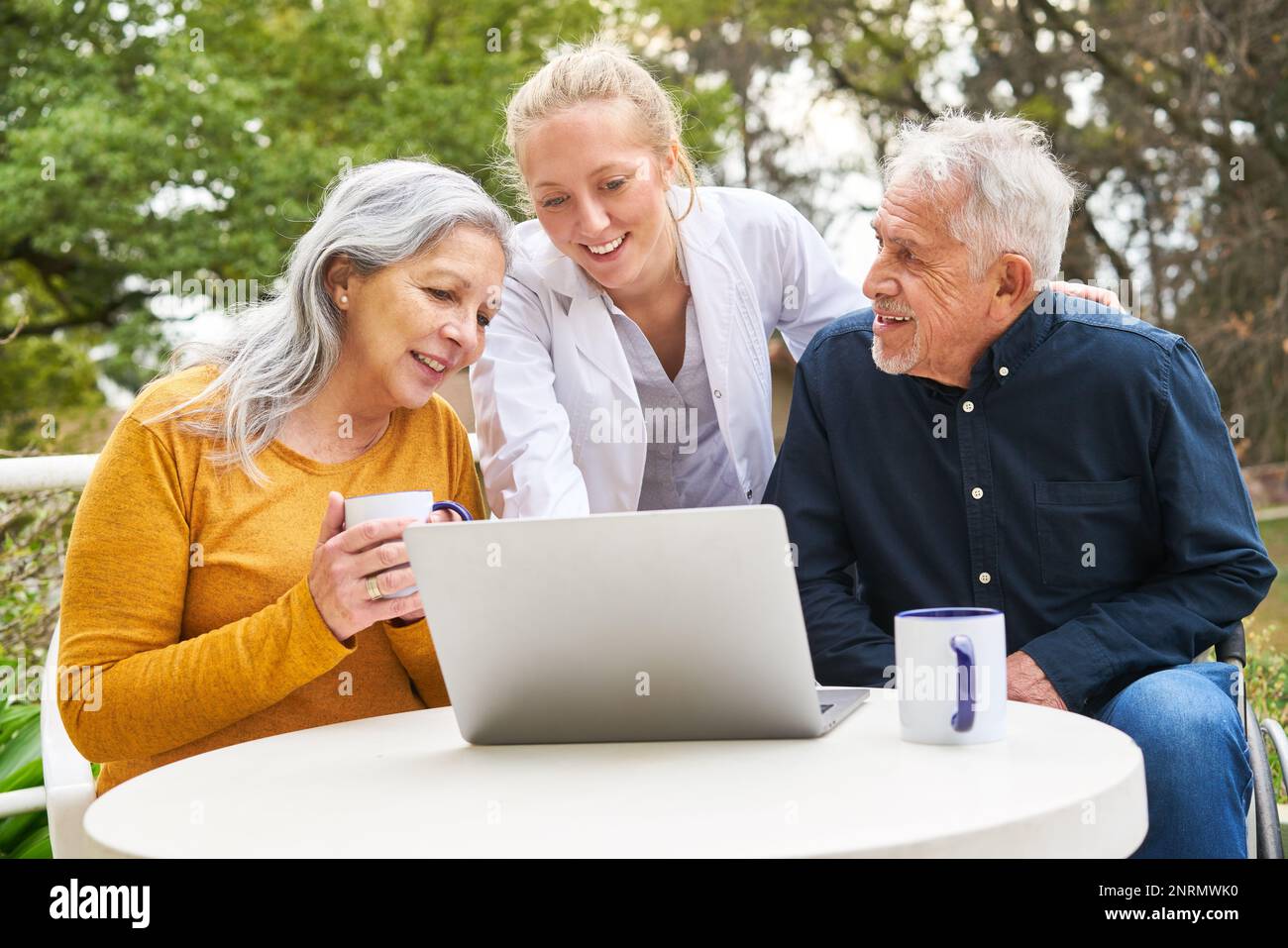 Una dottoressa che spiega la coppia anziana sul computer portatile mentre si ha un caffè a tavola nel giardino della casa di cura Foto Stock