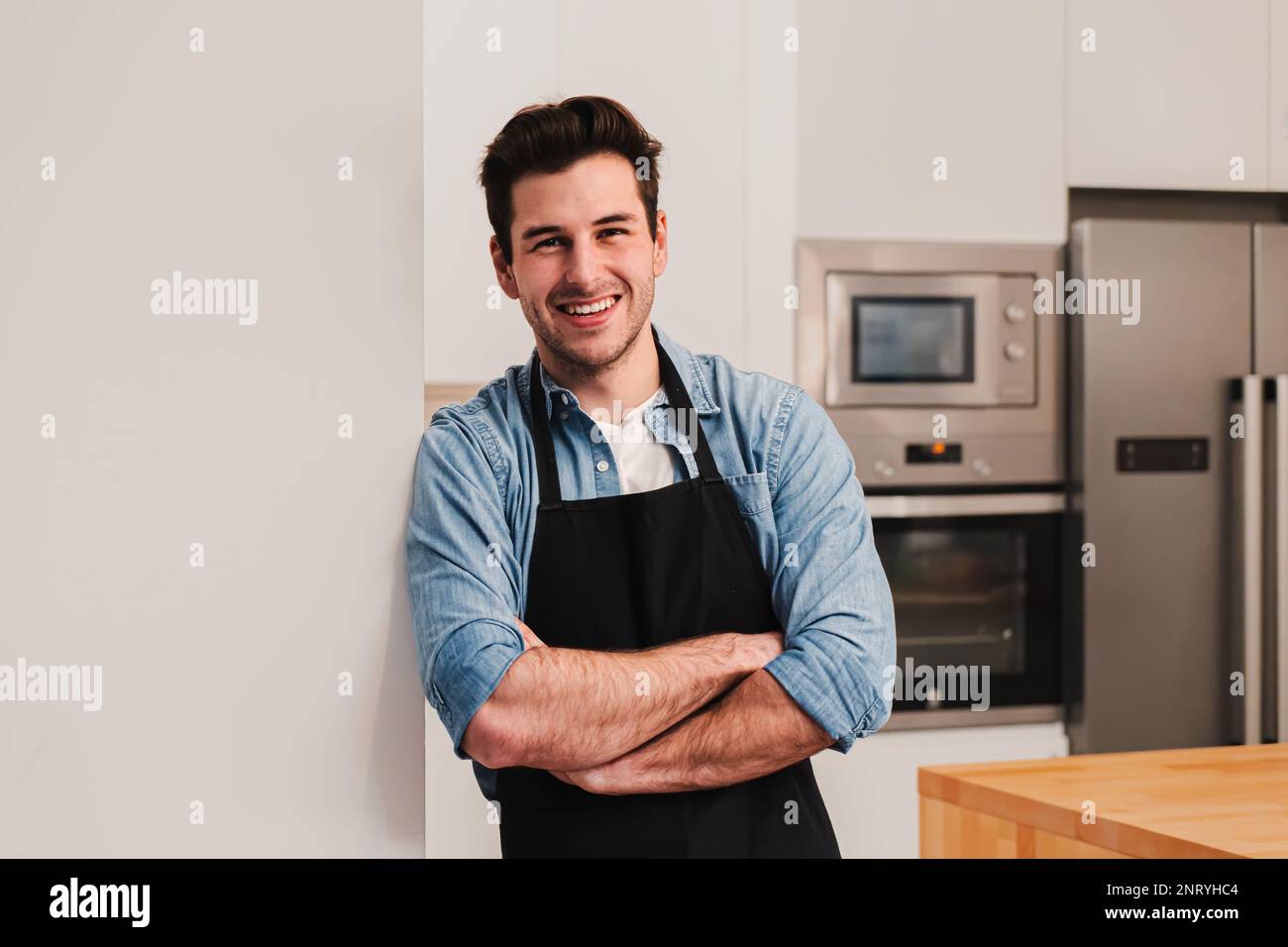 Attraente uomo caucasico che indossa un grembiule in piedi in cucina, sorridendo e incrociando le braccia guardando la macchina fotografica. Vista frontale di un bel maschio felice appoggiato a un muro a casa. Foto di alta qualità Foto Stock