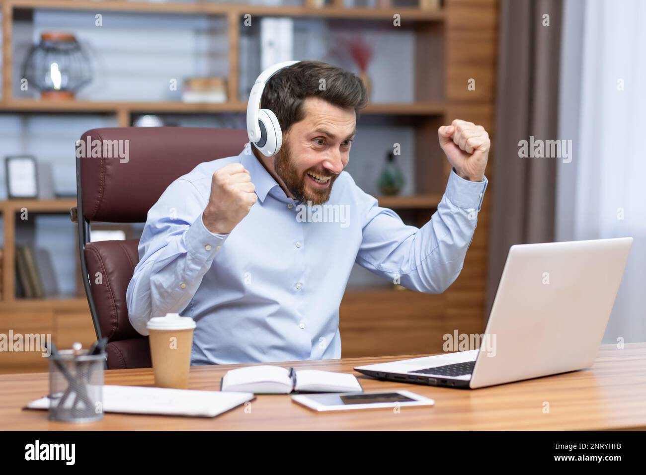 Uomo d'affari maturo in camicia e cuffie che guarda la partita sportiva sul posto di lavoro all'interno dell'ufficio, l'uomo che fa il tifo per divertirsi sul lavoro, guardando video sul laptop. Foto Stock