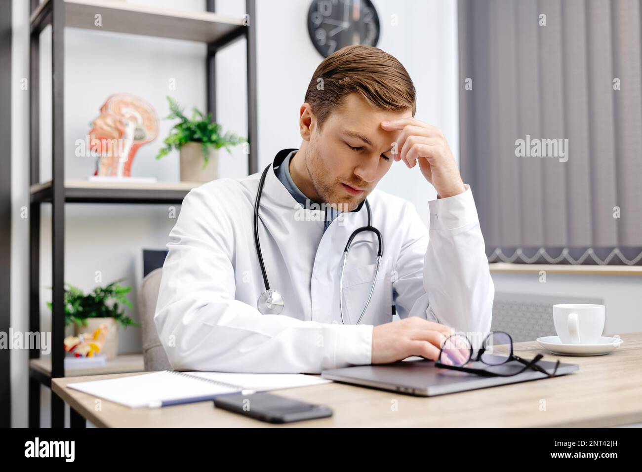 Bel dottore che indossa l'uniforme medica seduto sul suo posto di lavoro stanco tenendo la testa sensazione di stanchezza e mal di testa. Concetto di stress Foto Stock