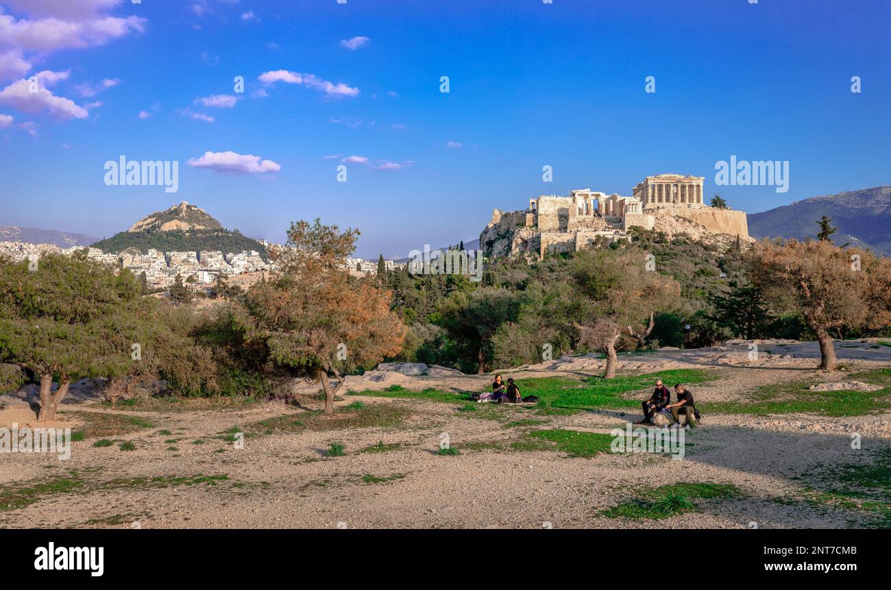 La gente incidentale sulla collina di Pnyx gode di un pomeriggio di sole, con l'Acropoli di Atene e il Monte Licabetto sullo sfondo. Atene, Grecia. Foto Stock