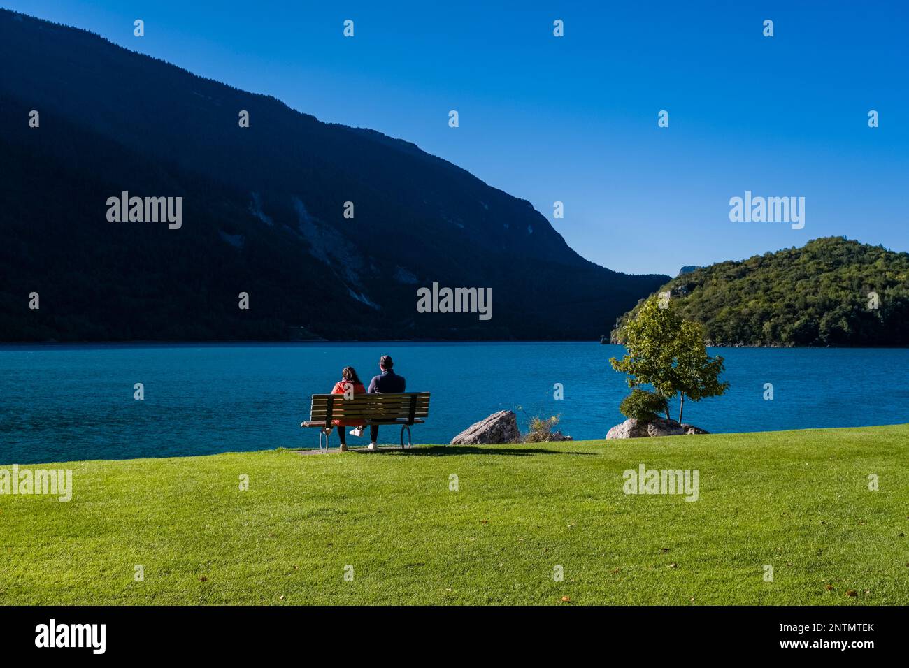 Due persone sedute su una panchina di legno sul lago di Molveno, pendici collinari e boschi in lontananza. Foto Stock