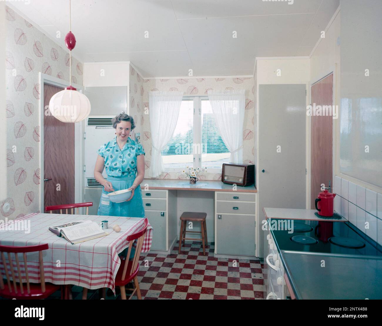 Ascoltatori radio in passato. Una donna in cucina sta frustando qualcosa in una ciotola al tavolo da cucina. Una radio è visibile sul banco di lavoro dalla finestra. Sulla parte anteriore della radio è presente una scala e quando si ruota la manopola accanto ad essa in modo che il puntatore si trovi al centro del nome di una stazione, la stazione era lì. Oltre alle trasmissioni FM, hanno anche ascoltato le radio straniere a onde lunghe. Svezia nel 1958 Foto Stock