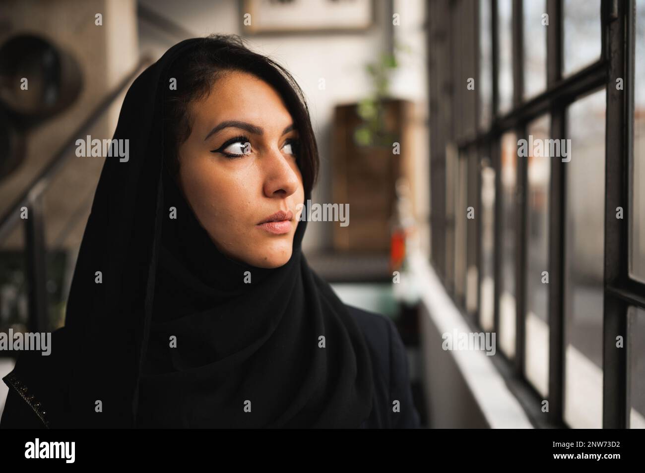 Primo piano di una giovane e attraente donna araba che indossa un velo nero. Profilo femmina che guarda fuori dalla finestra con faccia seria. Foto Stock