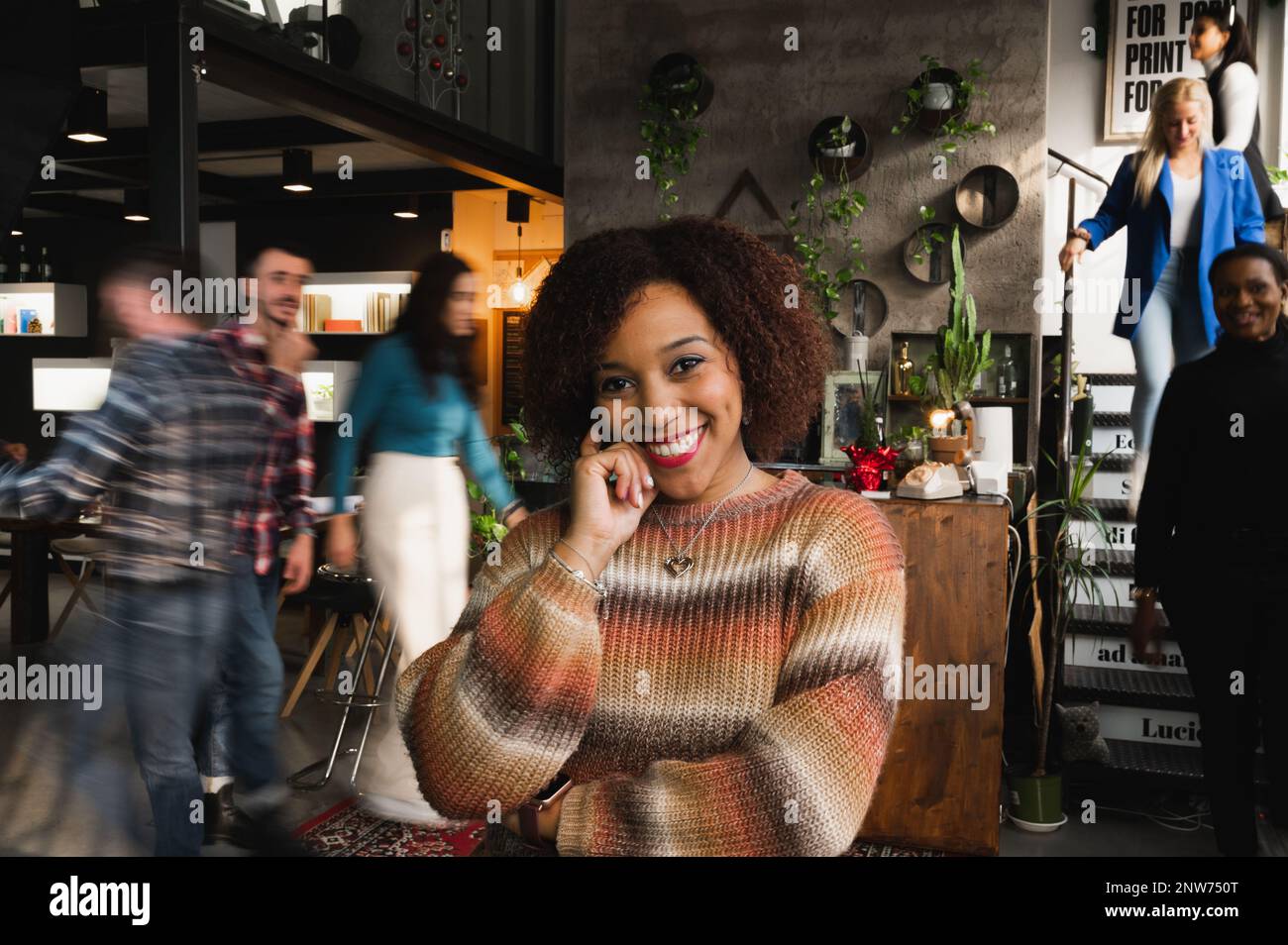 Ritratto di sorridente donna latina attraente guardando la macchina fotografica. Casual femmina in un luogo di lavoro occupato con persone sfocate a piedi. Foto Stock