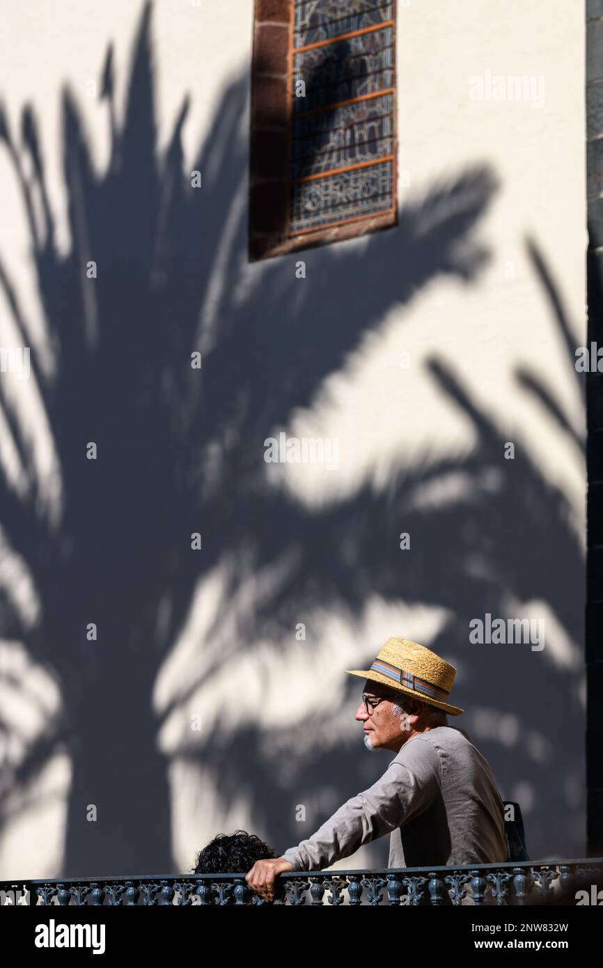 Un uomo con un cappello solare si trova accanto a una grande ombra di un albero di plam, gettato sul muro della Chiesa dell'Immacolata Concezione a la Laguna, Tenerife Foto Stock