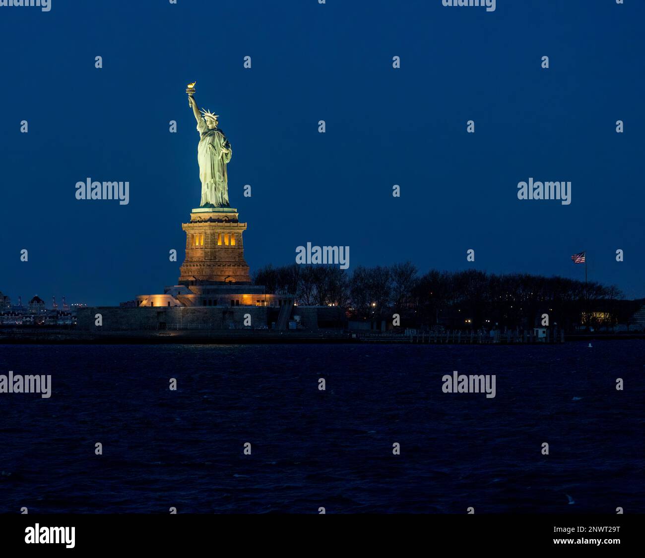 Profilo notturno della Statua della libertà dall'acqua con la bandiera degli Stati Uniti sullo sfondo Foto Stock