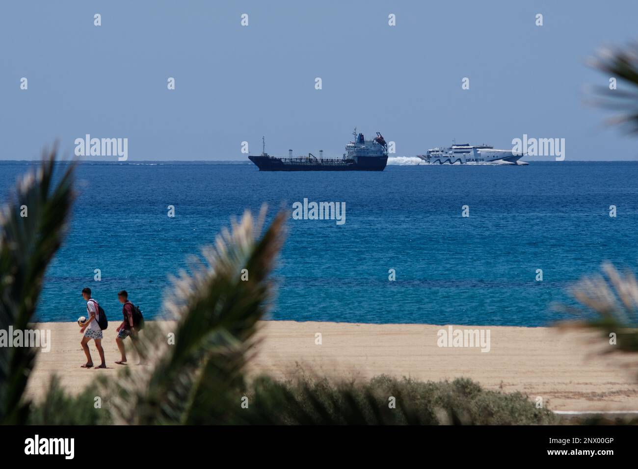 IOS, Grecia : 29 maggio 2021 : Vista dei turisti che camminano sulla spiaggia di Mylopotas in Grecia di iOS, una nave da carico e un traghetto ad alta velocità in background Foto Stock