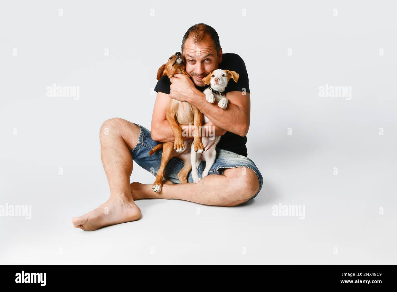 Un uomo seduto su uno sfondo bianco abbraccia affettuosamente i cuccioli del suo cane. Foto Stock