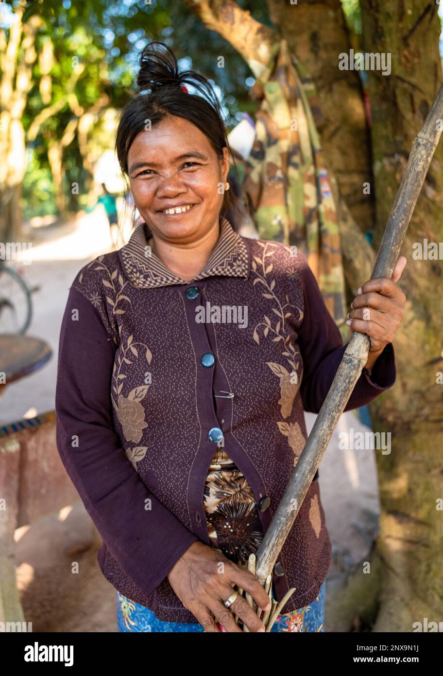 Una donna in un villaggio rurale nella provincia di Siem Reap, in Cambogia, tiene la sua scopa mentre lei spazza le foglie morte nel cortile ombreggiato fuori casa sua. Foto Stock