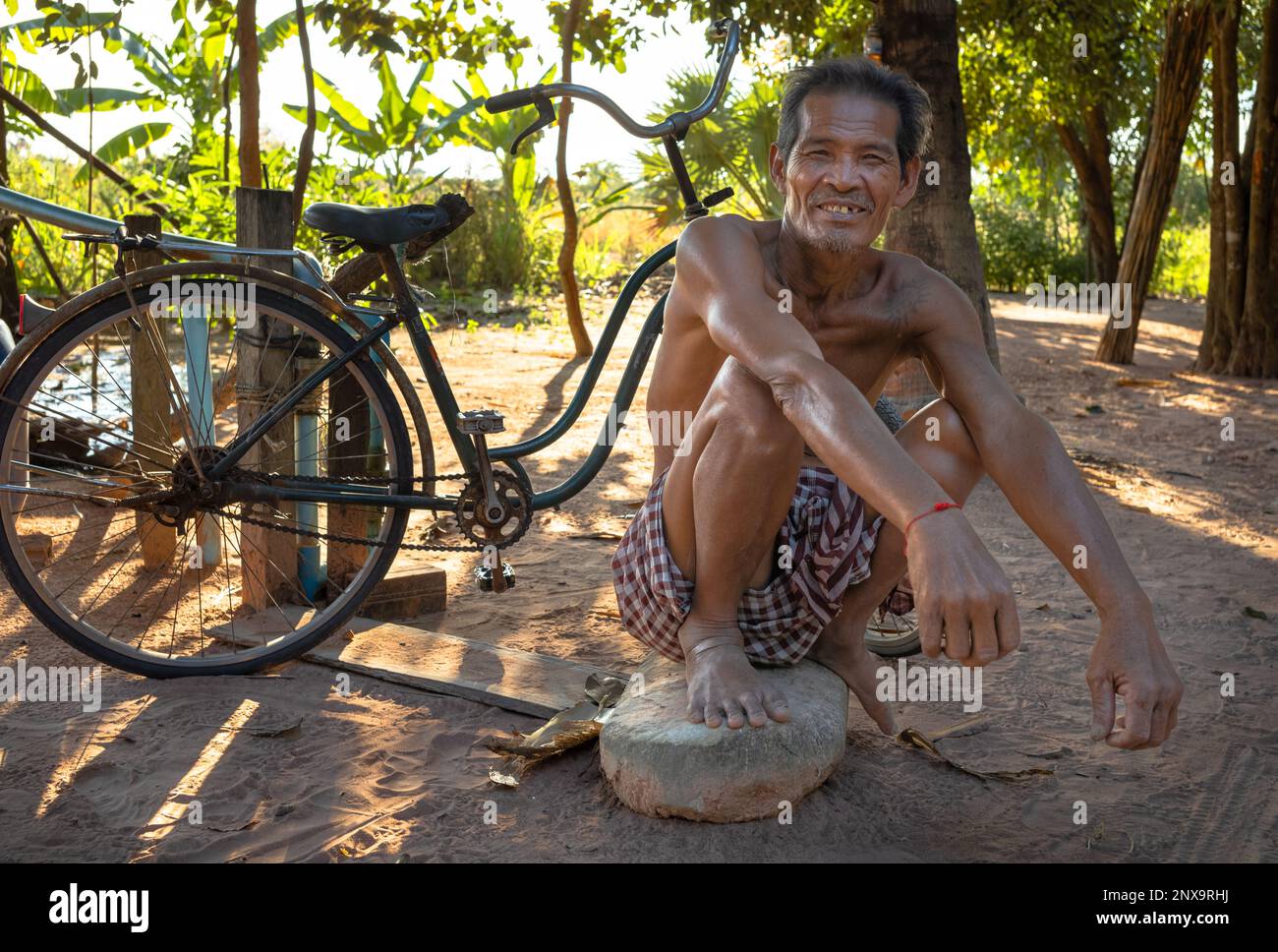 Un uomo di mezza età si schiaccia su una grande pietra accanto alla sua bicicletta in un villaggio vicino a Siem Reap in Cambogia. Foto Stock