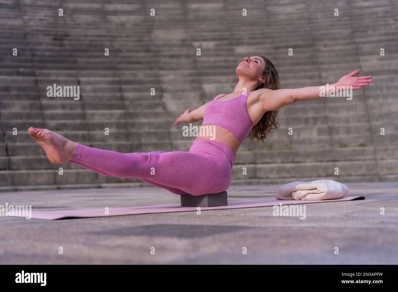 Una giovane donna caucasica seduta su un blocco di yoga con le gambe sollevate e le braccia tese Foto Stock
