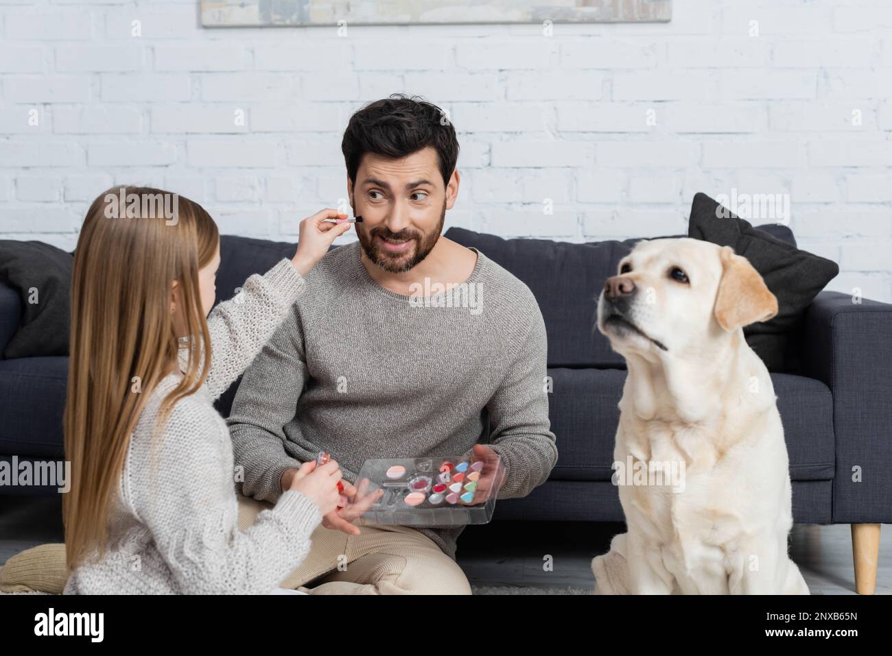 presen ragazza che tiene la tavolozza trucco mentre gioca con il papà sorridente vicino a labrador cane in soggiorno, immagine stock Foto Stock