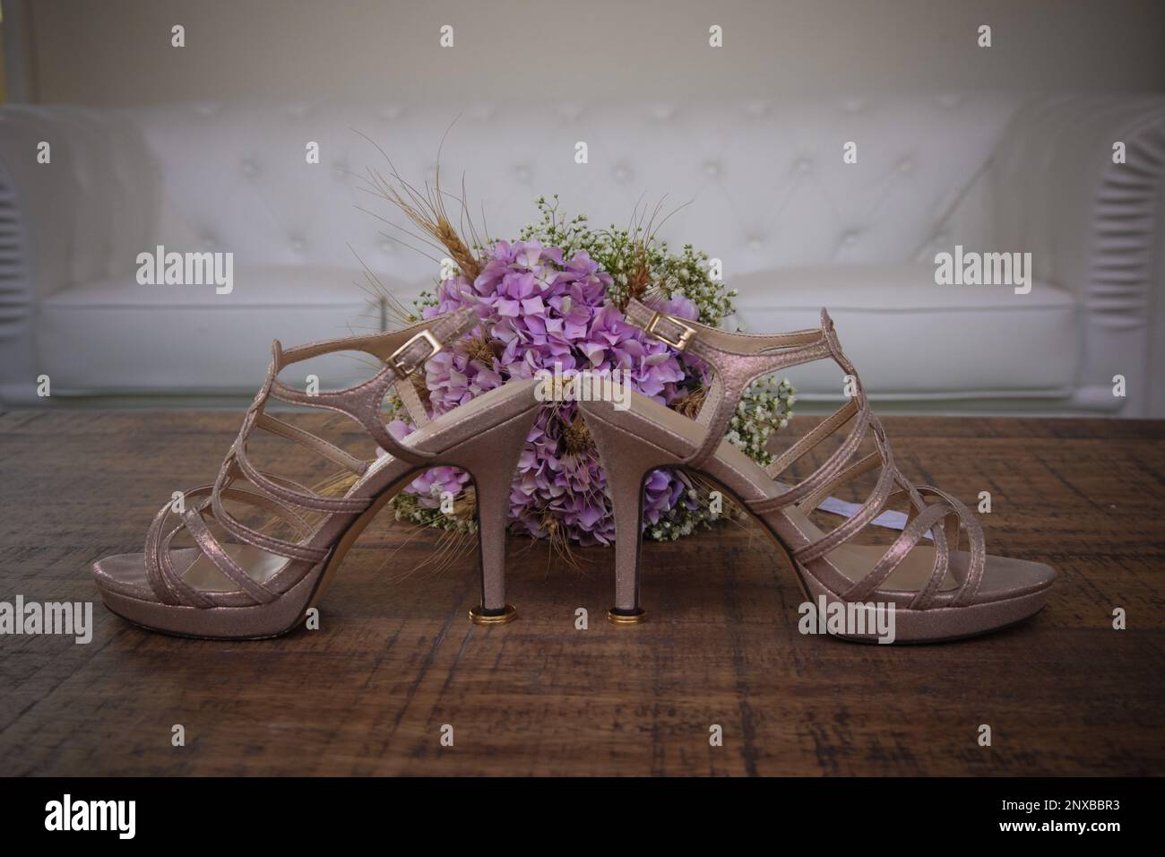 Primo piano di un paio di scarpe con tacco alto e bouquet da sposa su un tavolo di fronte a un divano Foto Stock