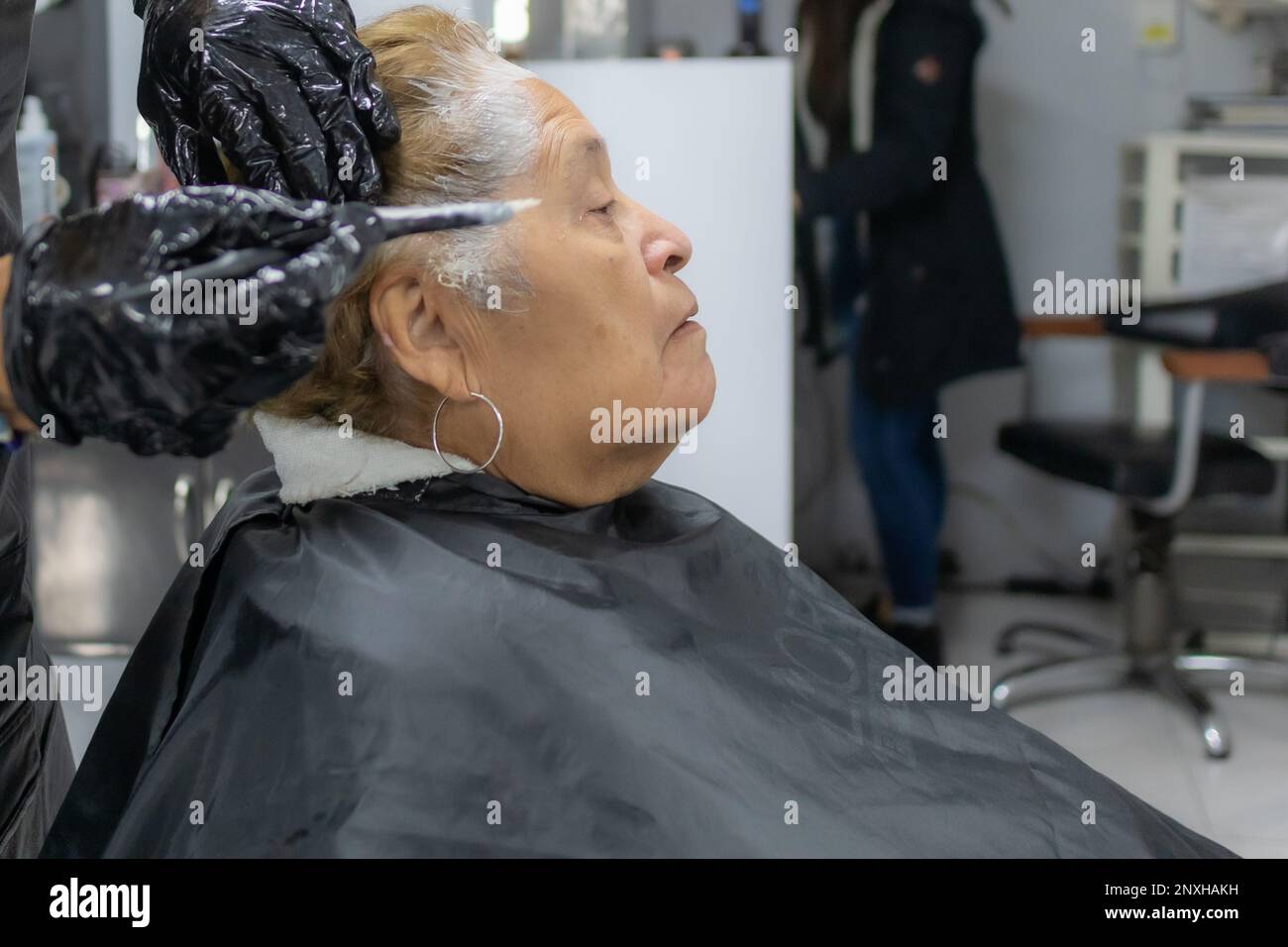 Stilista che inizia ad applicare colorante ai capelli corti di una donna anziana contro sfondo sfocato, espressione seria, vista laterale. Moda antica e bellezza c Foto Stock