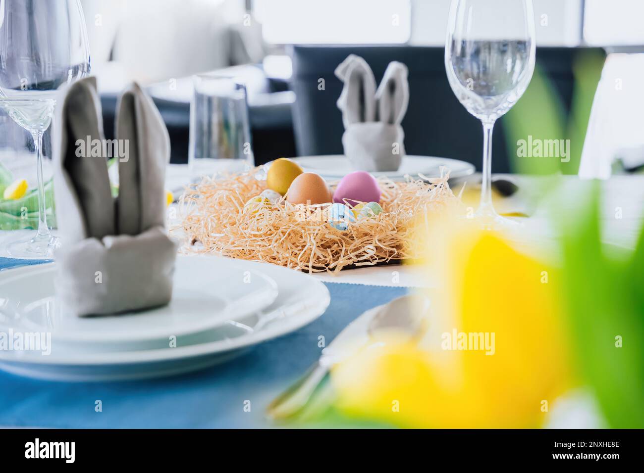 Bella cornice di Pasqua tavolo con arredamento festivo al coperto. Tovagliolo a forma di orecchie di coniglio, uova colorate in nido, tulipani gialli freschi in vaso. Sunligh Foto Stock