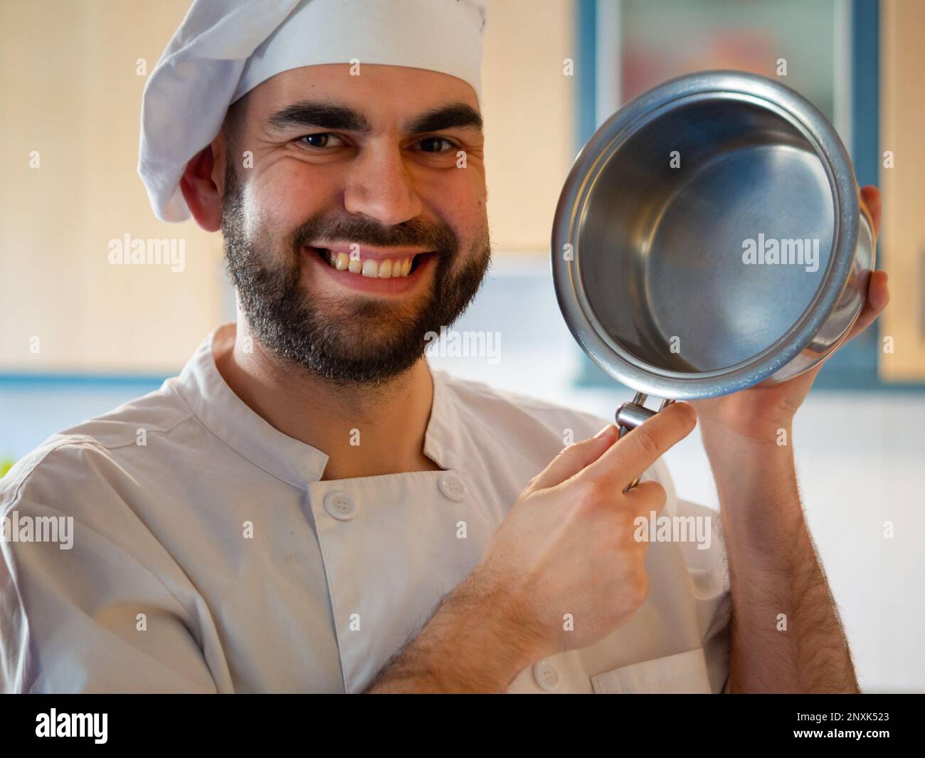 Ritratto di un giovane chef bearded sorridente mentre tiene una pentola in acciaio inossidabile guardando la macchina fotografica Foto Stock