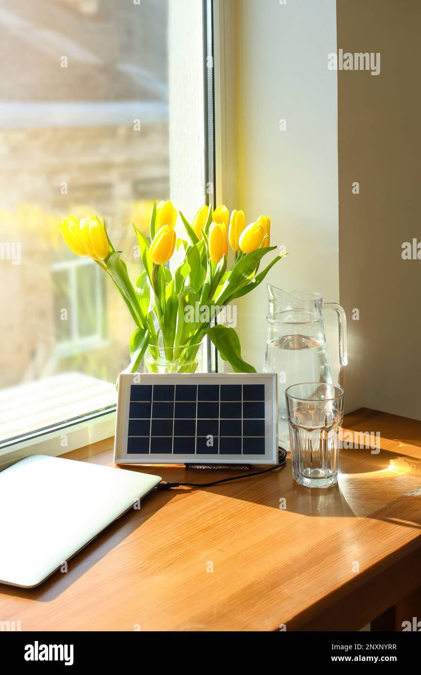 Portatile pannello solare di carica computer portatile, caraffa d'acqua e vaso con tulipani sul tavolo vicino alla finestra Foto Stock
