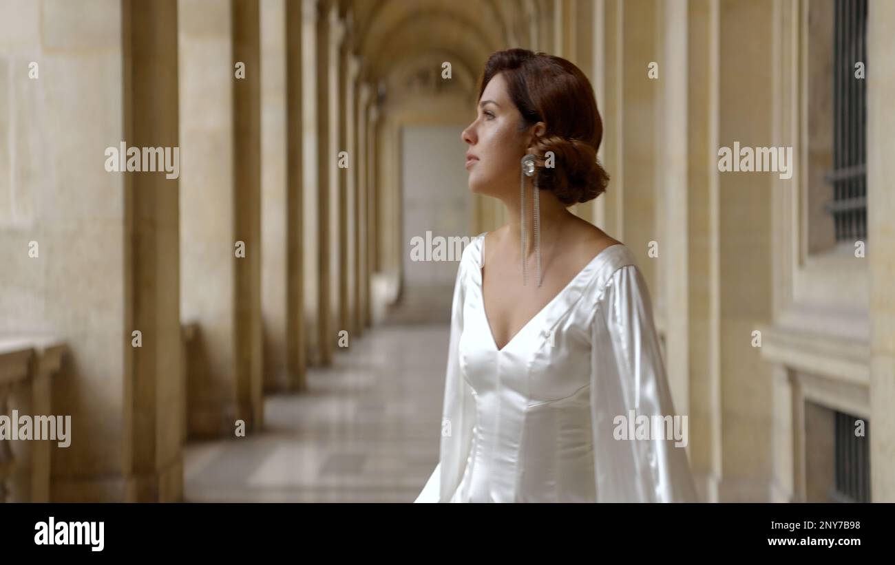 Donna in romantico abito di seta bianca guardando in lontananza sullo sfondo di un antico edificio. Azione. Donna elegante con orecchino lungo Foto Stock