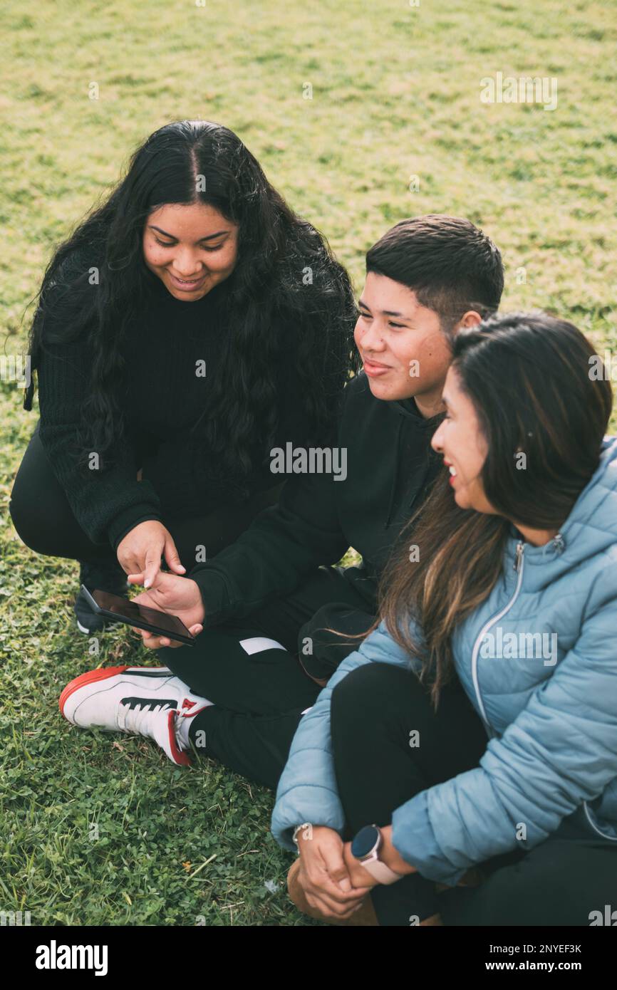 Tre individui di etnia ispanica-latina seduti sul verde lussureggiante di un parco, con dispositivo intelligente. Espressioni rilassate e sorridenti sul loro Foto Stock