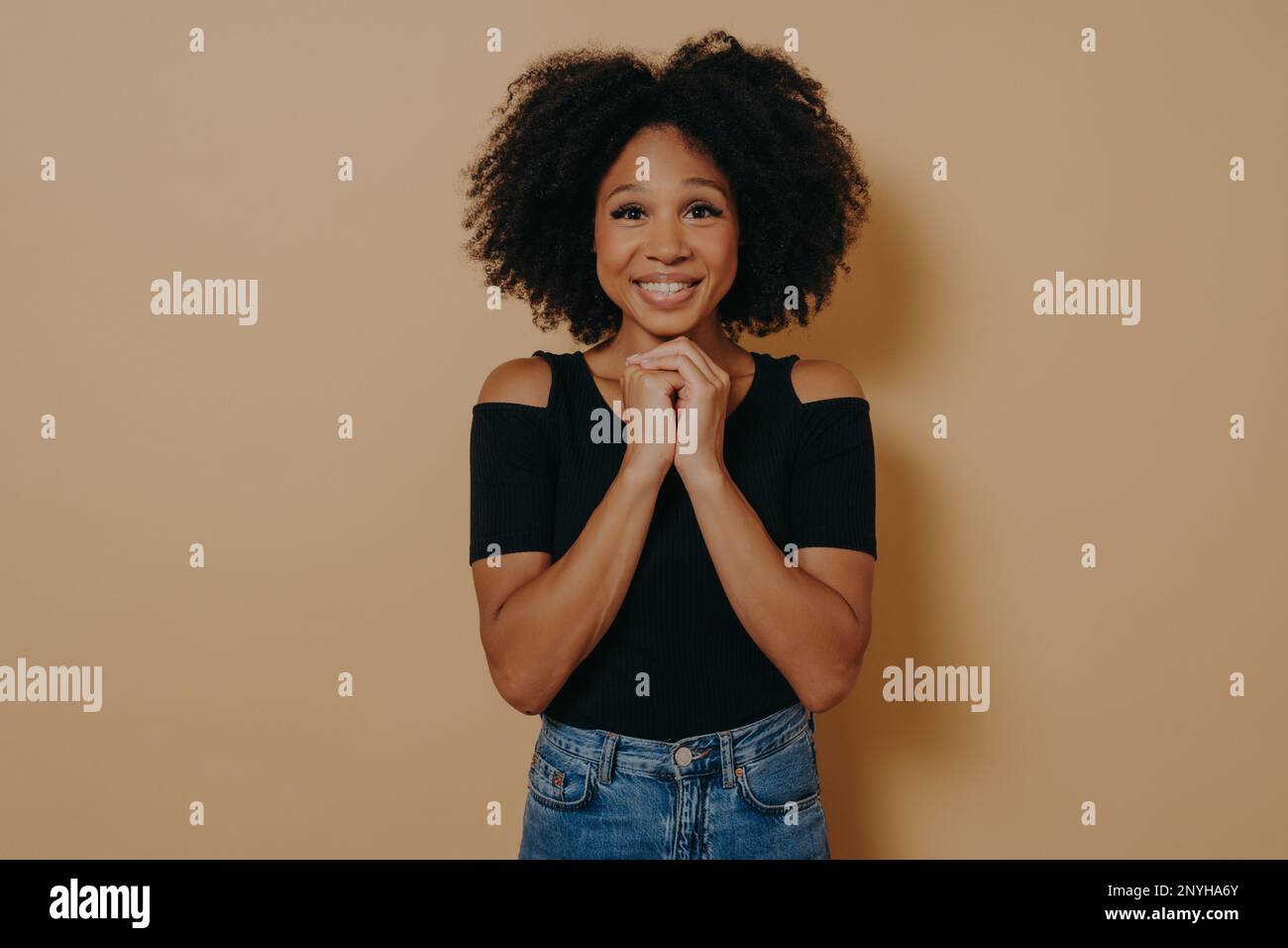 Giovane donna di razza mista che tiene le mani in gesto di preghiera, sorridente con gli occhi aperti isolato su sfondo beige scuro con spazio copia. Signora africana pos Foto Stock