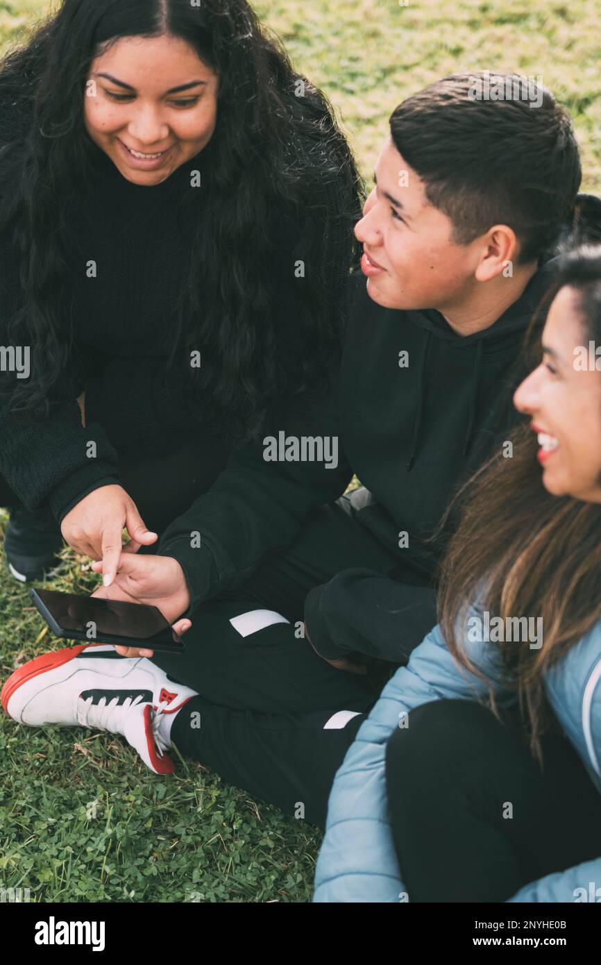 Tre individui di etnia ispanica-latina seduti sul verde lussureggiante di un parco, con dispositivo intelligente. Rilassato e sorridente famiglia ispanica spendere Foto Stock