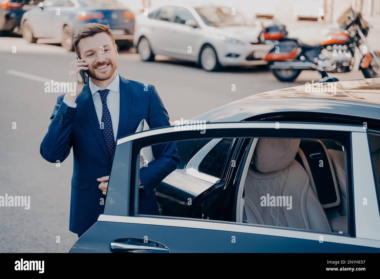Un dirigente sorridente e sicuro vestito formale blu che lascia la limousine nera dopo essere arrivato al suo posto di lavoro, parlando al telefono, tenendo il giornale w Foto Stock