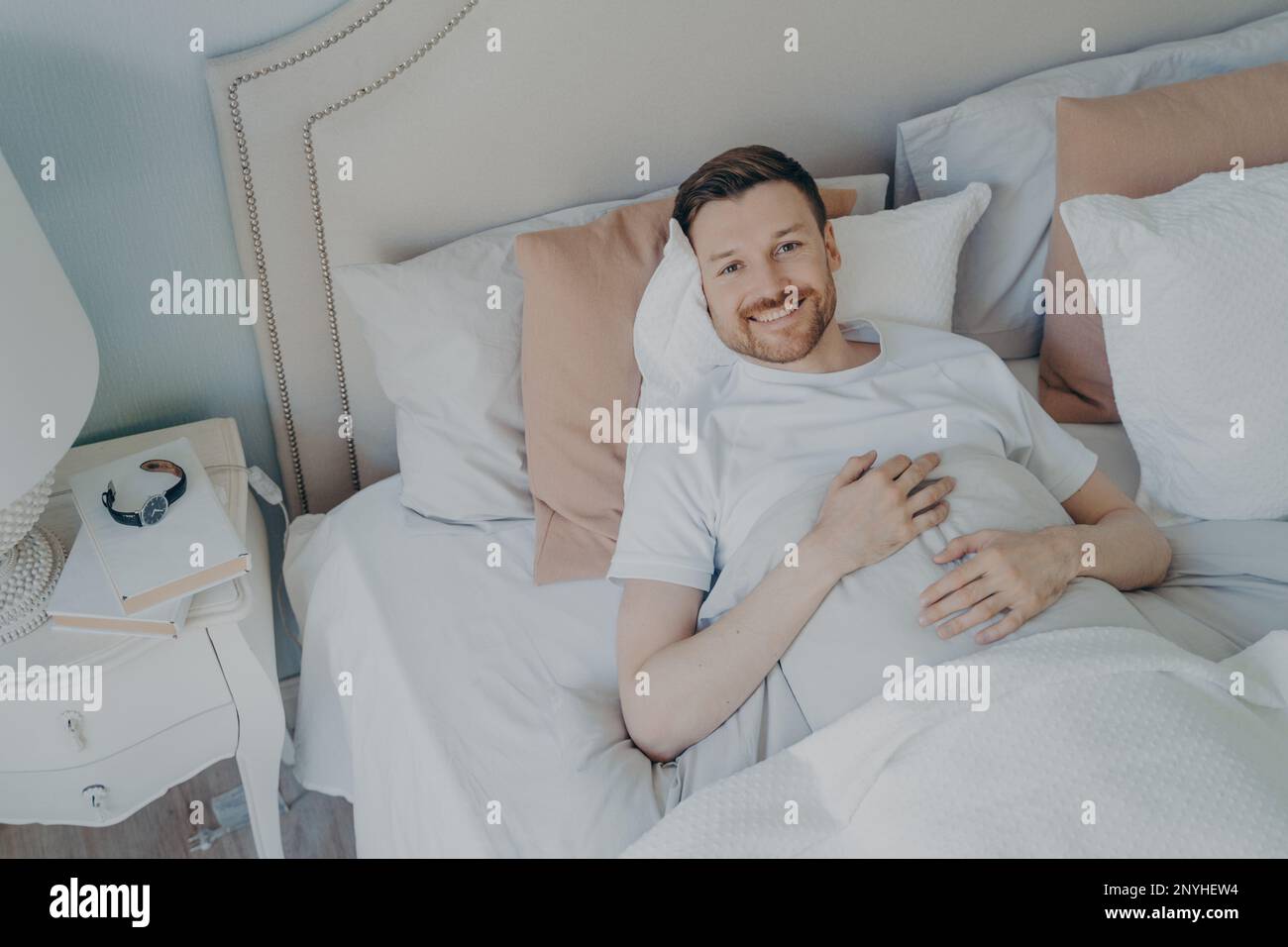 Giovane uomo ben riposato in maglietta bianca svegliarsi al mattino con sorriso, felice dopo aver dormito abbastanza, sdraiato in comodo letto mentre coperto di bl Foto Stock