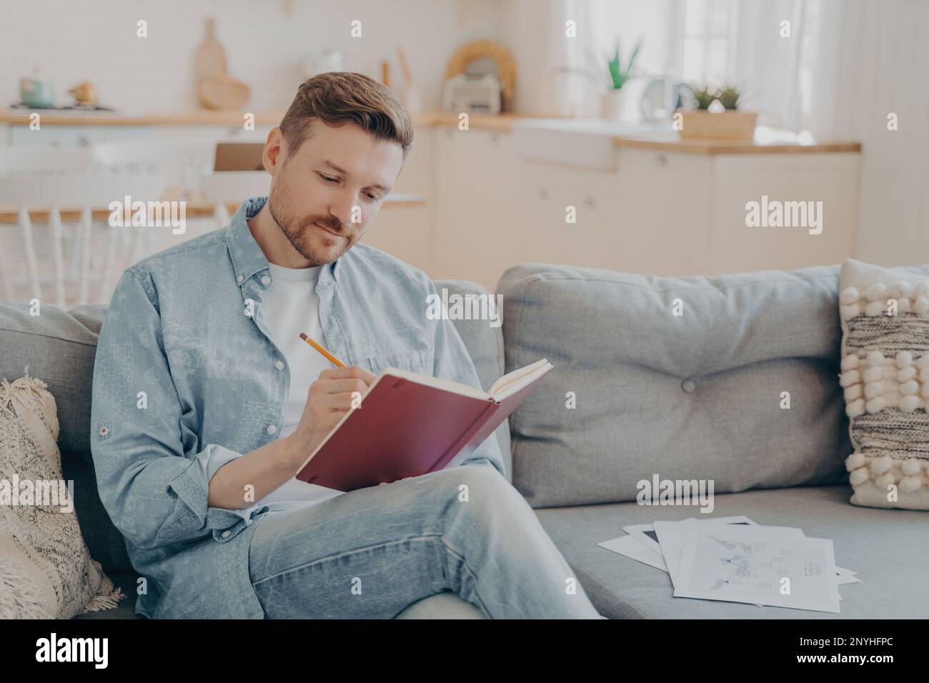 Occupato giovane uomo vestito in abiti casual scrivendo in taccuino rosso con matita mentre si siede su comodo divano con gambe incrociate, guardando concentrato, ki Foto Stock