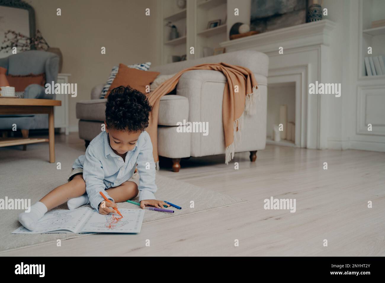 Piccolo ragazzo di razza mista disegno in libro da colorare con un sacco di pennarello diverso mentre si siede da solo sul pavimento in mezzo al soggiorno a casa. Foto Stock