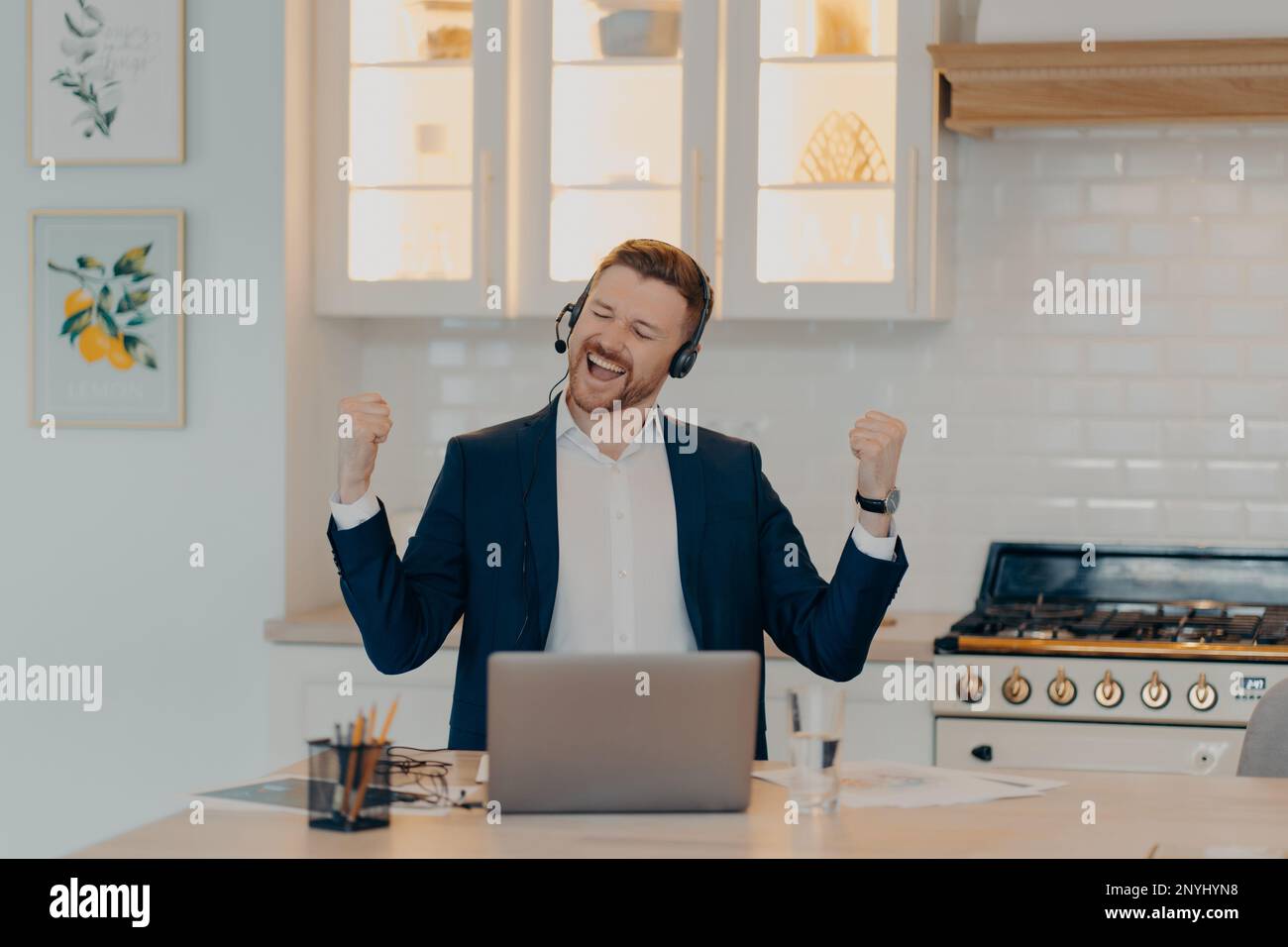 Uomo d'affari sorridente e con cuffie che raggiunge gli obiettivi aziendali al lavoro e festeggia il successo mentre si siede al tavolo con un computer portatile in cucina, Foto Stock