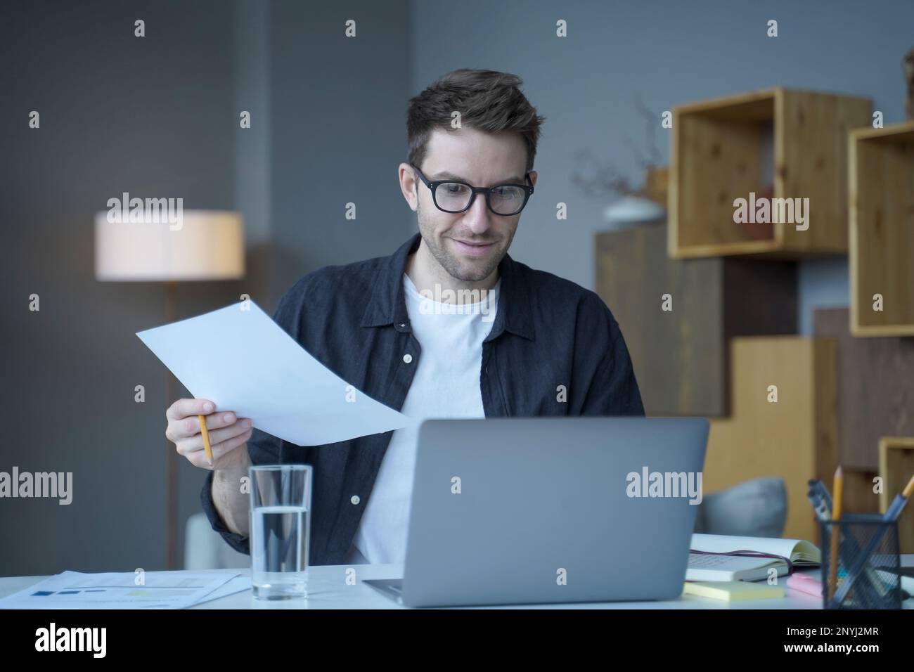 Sicuro uomo positivo consigliere finanziario in eyewear funziona in remoto on-line a casa moderna ufficio. Dipendente sorridente dell'azienda che tiene documenti cartacei, c Foto Stock