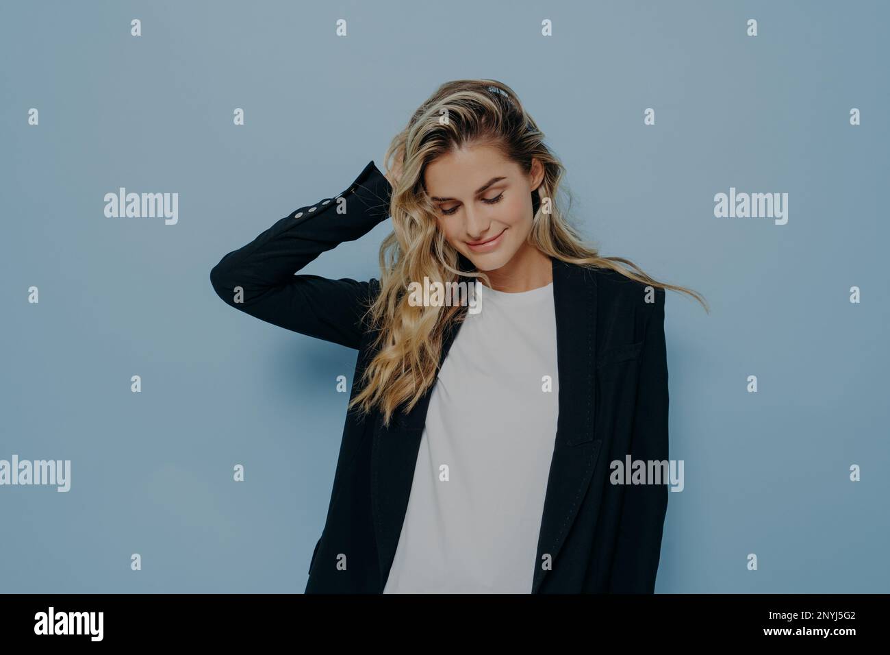 Bella donna sorridente con capelli biondi tinti sentirsi felice, indossando biondo nero su maglietta bianca mentre si è in piedi isolato su sfondo blu, Foto Stock