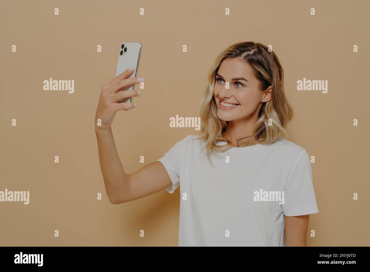 Giovane donna bionda sorridente che usa lo smartphone mentre videochiama con la famiglia o l'amico, vestita di maglietta bianca, guardando la fotocamera mobile e gli sili Foto Stock